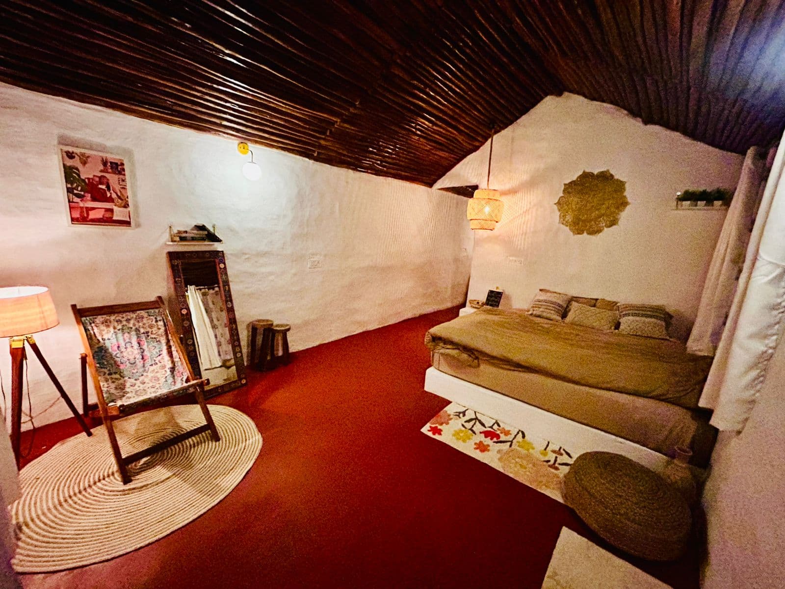 A mud house bedroom with whitewashed walls, a wooden ceiling, a jute rug and a folding chair